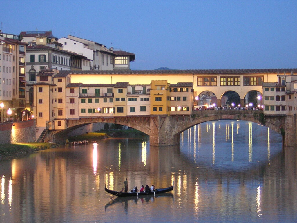 Photograph of Florence's iconic Ponte Vecchio, the Old Bridge. A lower stone section with arches is covered with tiny houses in various shades of golden stucco, with little square widows with green or red shutters. Along the upper portion going across above the roofs of the tiny buildings is a long yellow corridor, matching what we saw wrap around the tower. The picturesque combination is photographed in twilight, with lights shimmering on the deep blue water. In the river below, a totally inappropriate gondola full of tourists is looking up at the bridge (Florence did not have gondolas, only Venice did, this is very silly, but very pretty!)