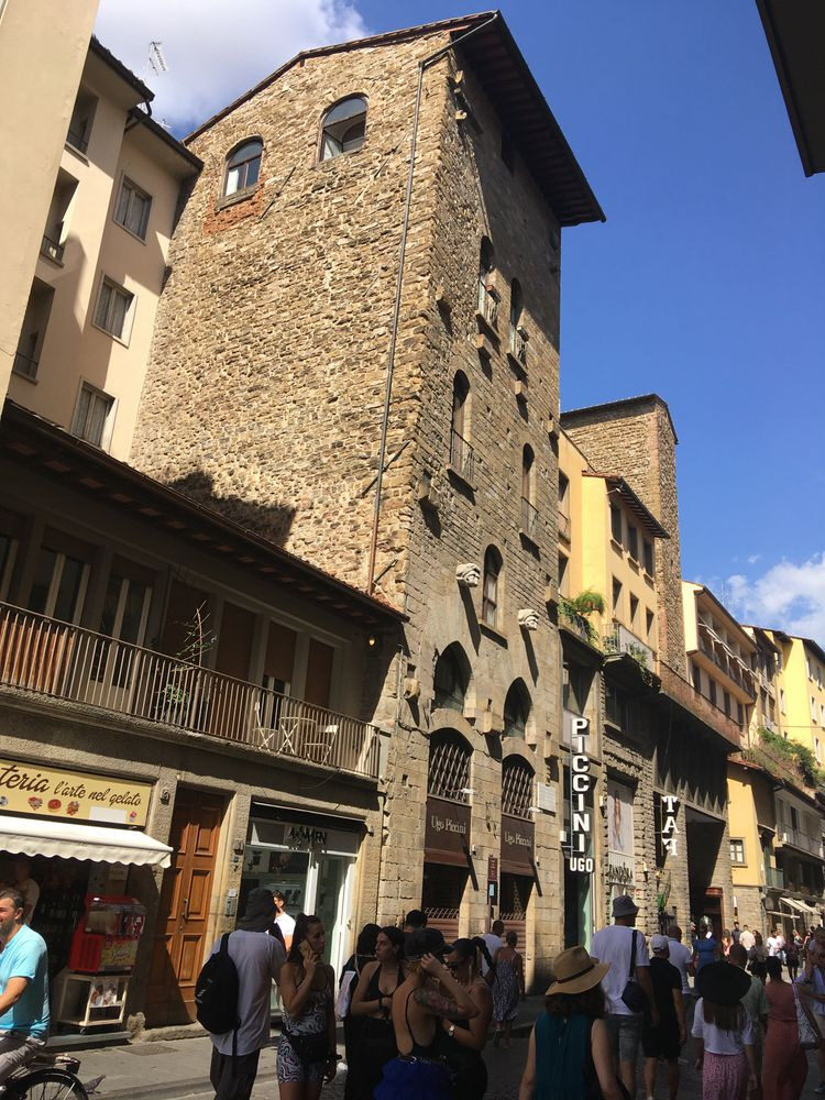 A photo of a street in Florence. Many tourists walk along and the buildings are all sho ps and eateries. In the center, conspicuous between buildings of yellow or beige stucco, is one building made of crude - looking yellowish stone, very rough and undecorated, with few windows and all small compared to its neighbors. A couple doors down, a sec ond conspicuous stone section like this sticks up, also strangely blank and rough amid its yellow neighbors. Both stop about half a story above the roofs of the three - story buildings on either side of them.